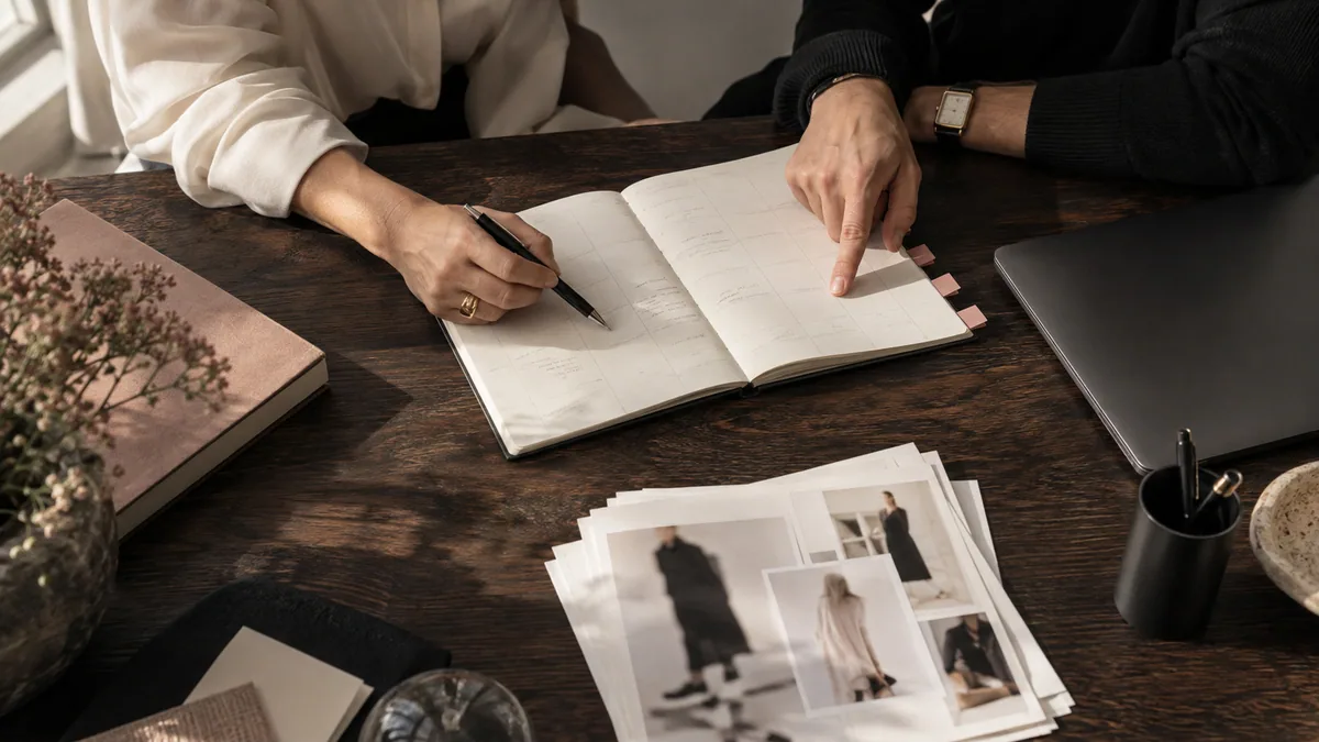 Two people review a paper content schedule and printed contact sheets beside a laptop at a work table.